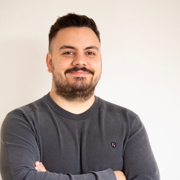 Ein Mann mit kurzem Bart steht mit verschränkten Armen und lächelt freundlich.