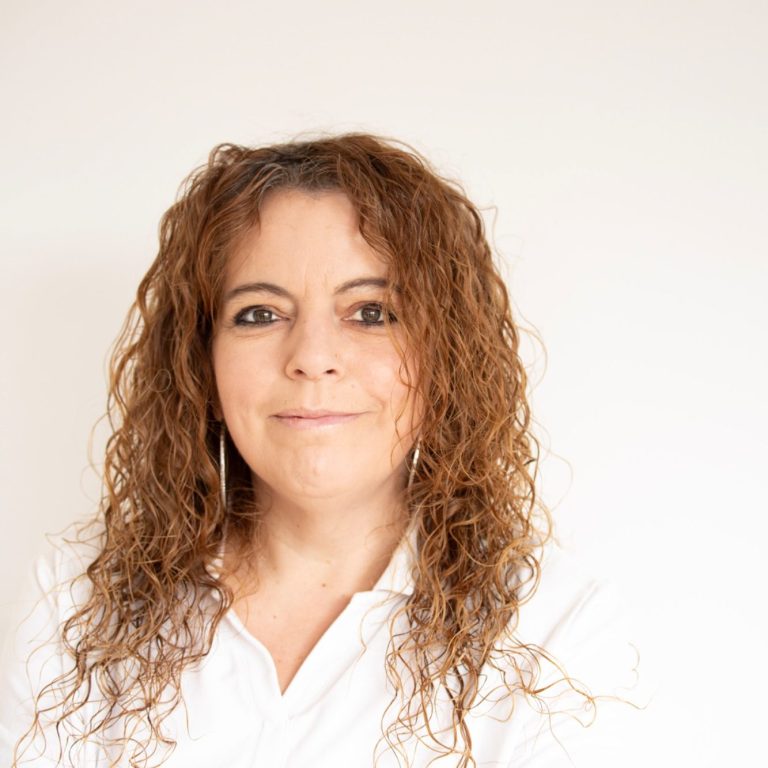 Lächelnde Frau mit lockigem, schulterlangem Haar in einem weißen Hemd vor hellem Hintergrund.